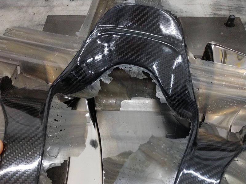 Image shows the head and neck safety device just removed from its tool in a production environment. The composite bladder molded structure has resin flashing along the edges of the hardware where the tool halves met. The surface of the hardware is a high gloss carbon fiber weave.